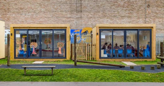 Two outdoor classroom garden rooms with small group of children sat on blue chairs in right building. Playground equipment in the foreground.