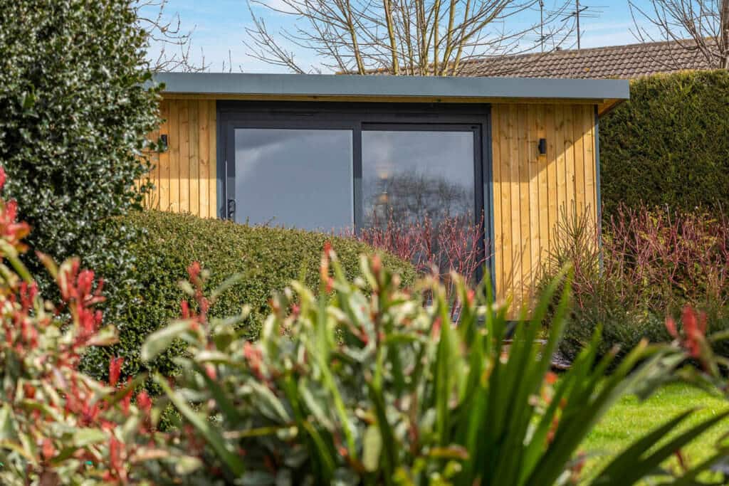 Redwood clad garden studio with sliding graphite doors peeking out behind garden shrubs.