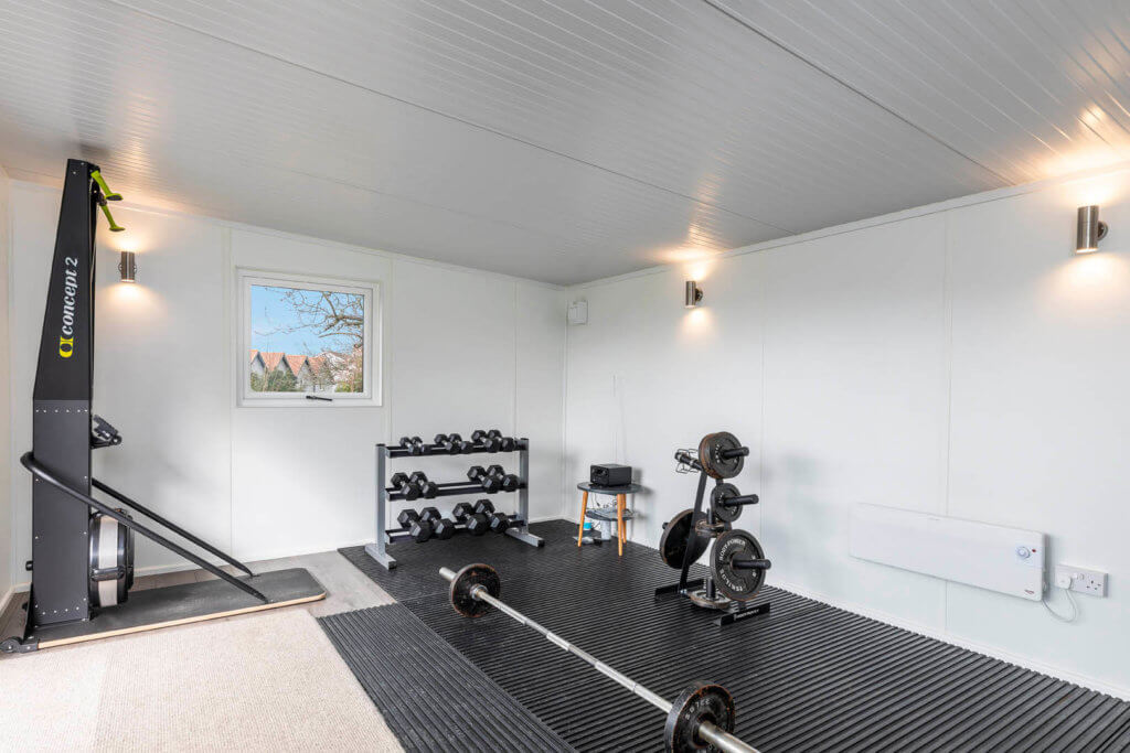 Interior of garden gym with carpet and black matting floor with weights and dumbbell rack.