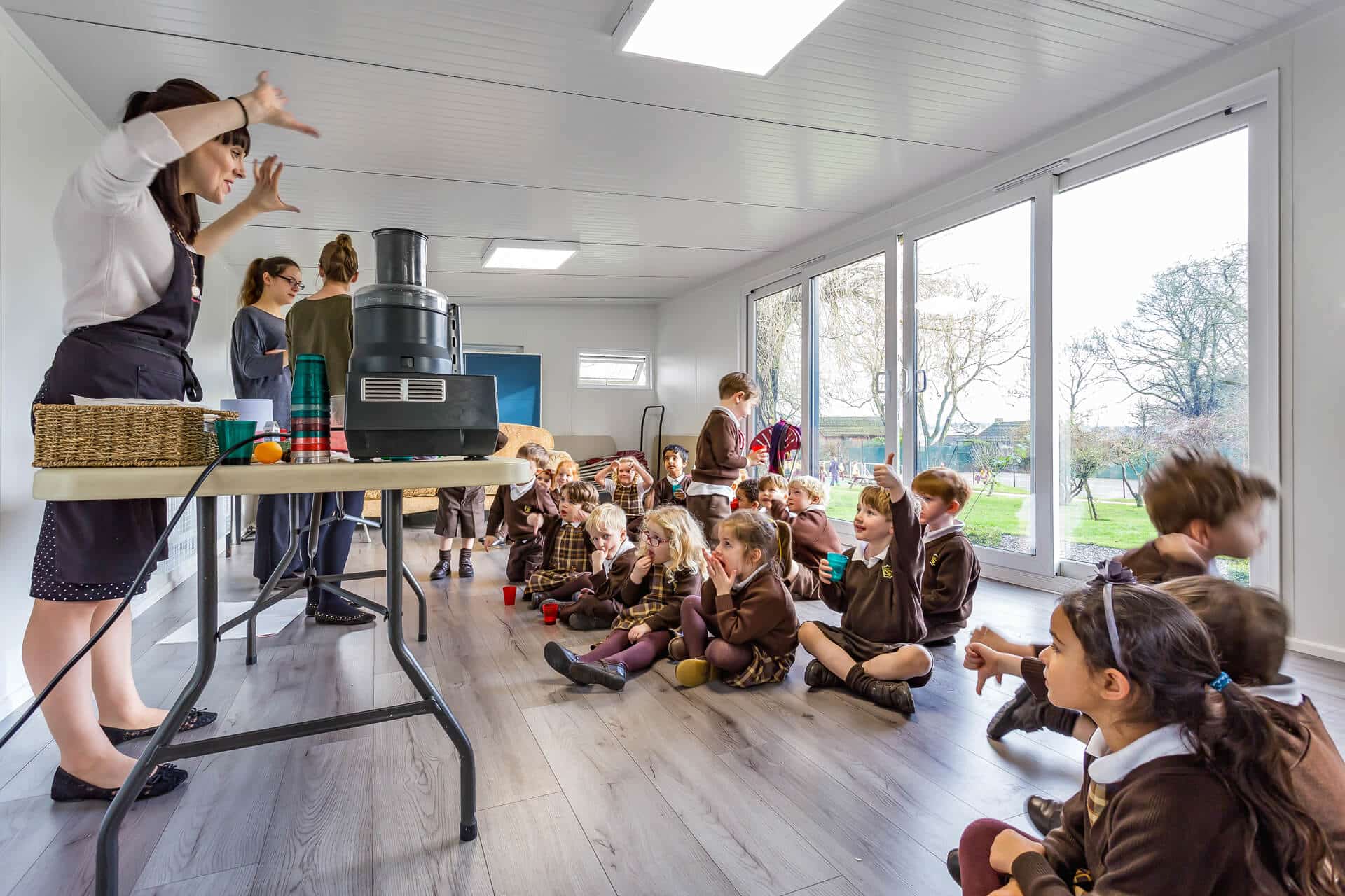 Interior side view of outdoor classroom building with light oak flooring. Female teacher stood at front engaging a group of primary school children in brown uniform.