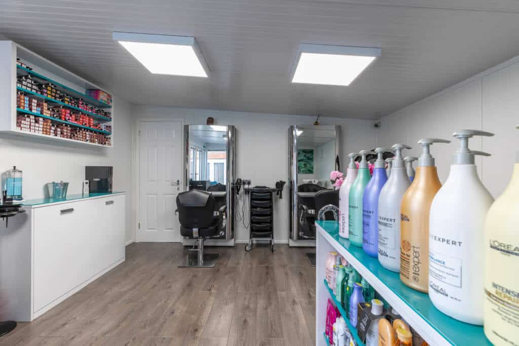 Multi coloured shampoo and conditioner bottles on shelf in the foreground. Salon chairs and full length mirrors in the background.