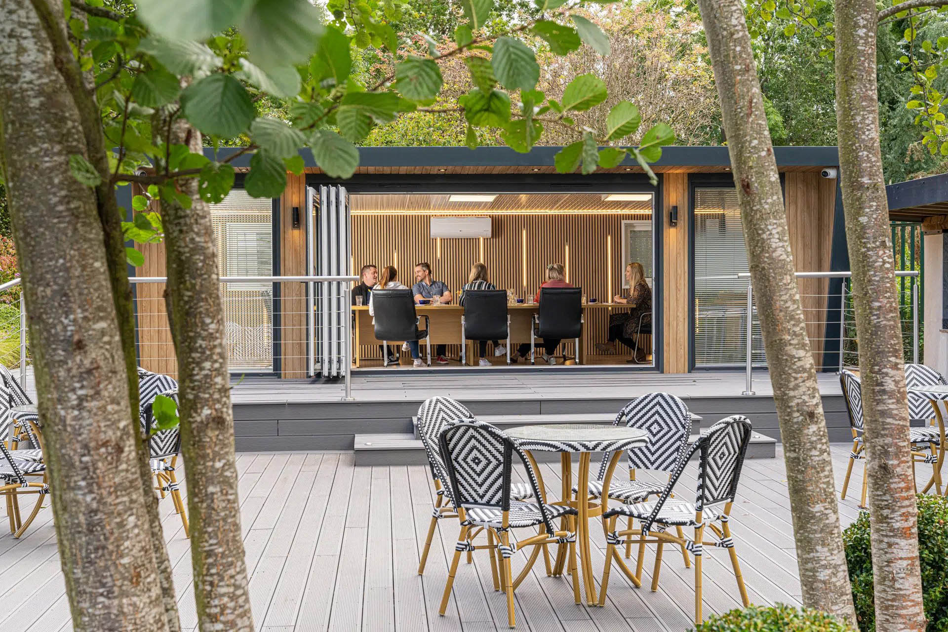 Exterior of a boardroom with people inside having a meeting. The building is on a raised patio and there is garden furniture outside
