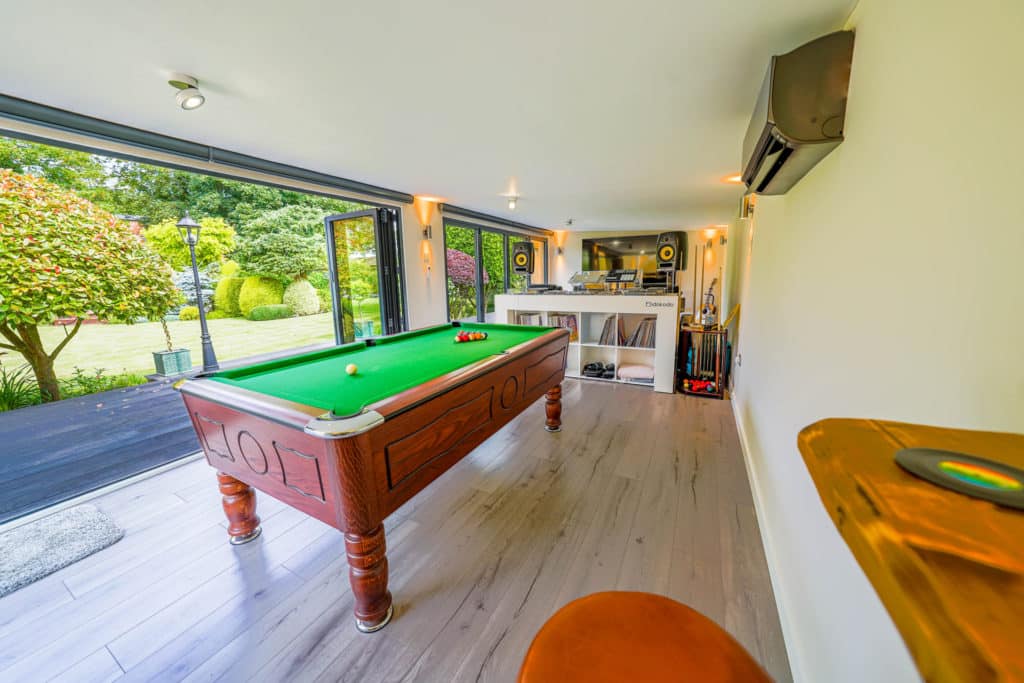 Interior of a man cave with two sets of bi-fold doors, a pool table in the centre, a DJ booth behind it and a flat screen TV in the bacground