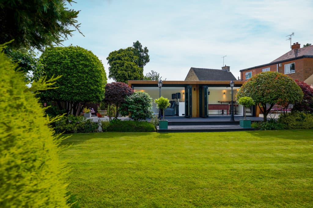 Exterior of a man cave in a landscaped garden with two bi-fold doors open looking into a home cinema and a pool table