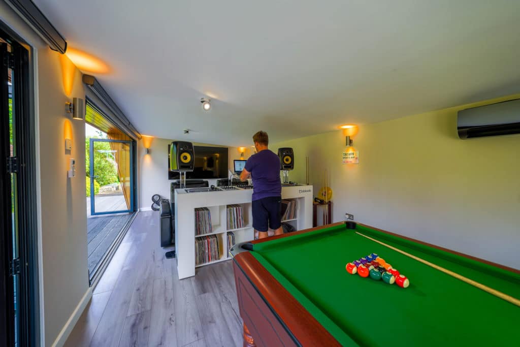 Pool table in the foreground with a cue a pool balls, a man in a purple t-shirt and blue shorts standing at a DJ booth behind that and a large flat screen TV in the background in a man cave