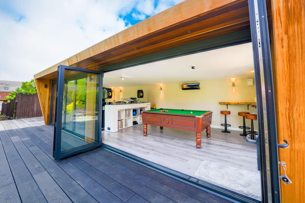 Exterior of a man cave on a decked patio with its bi-fold doors open, looking into an entertainment space