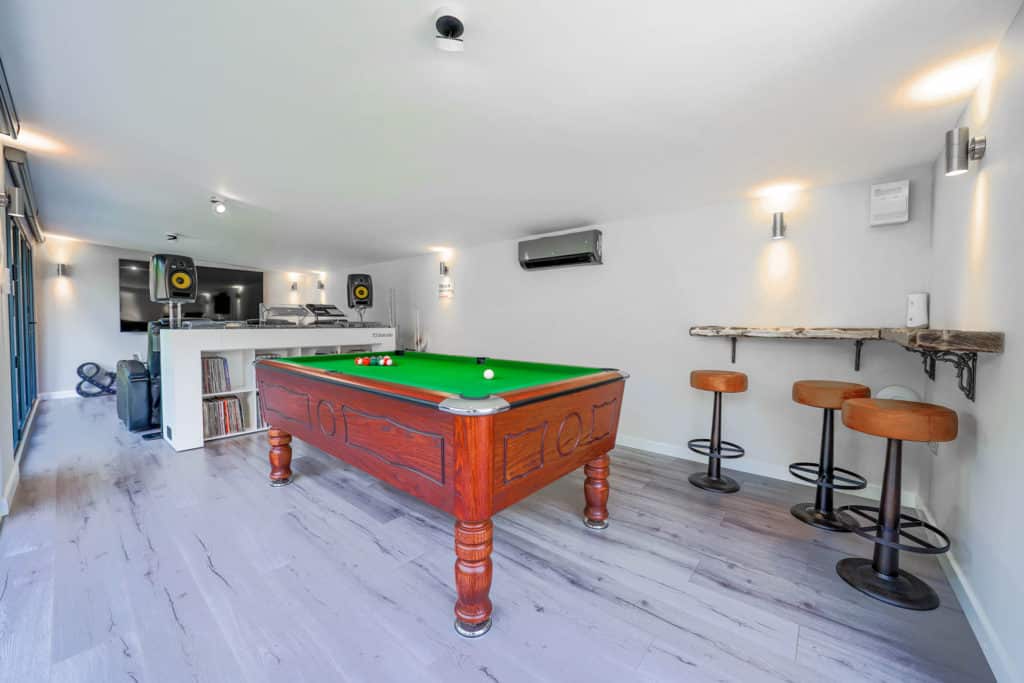 Interior of a man cave with a bar and stools in the right corner, a pool table next to it, a DJ booth behind that with large speakers and a large flat screen TV on the left back wall