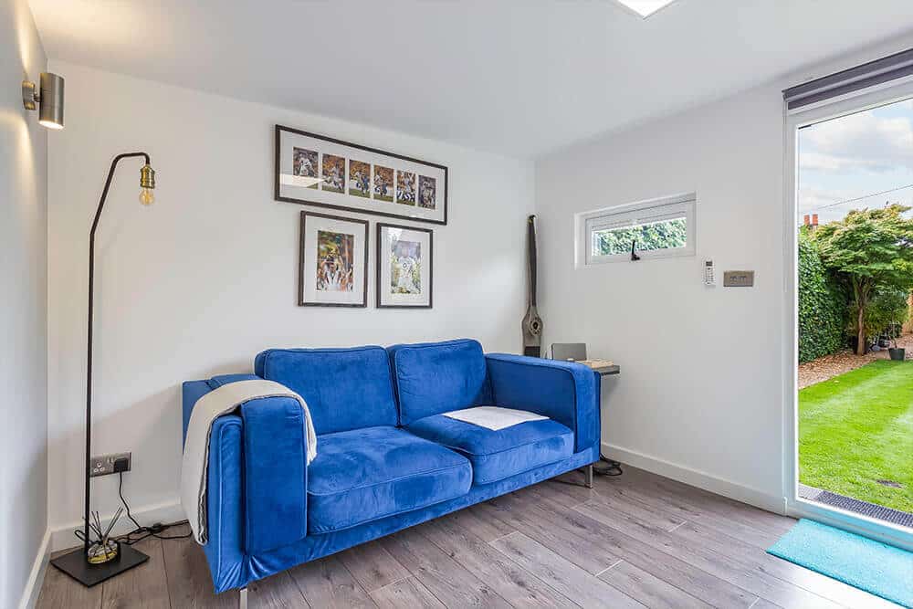 Electric blue sofa in garden room with framed rugby images on the wall behind. Modern black floor lamp tot he left.