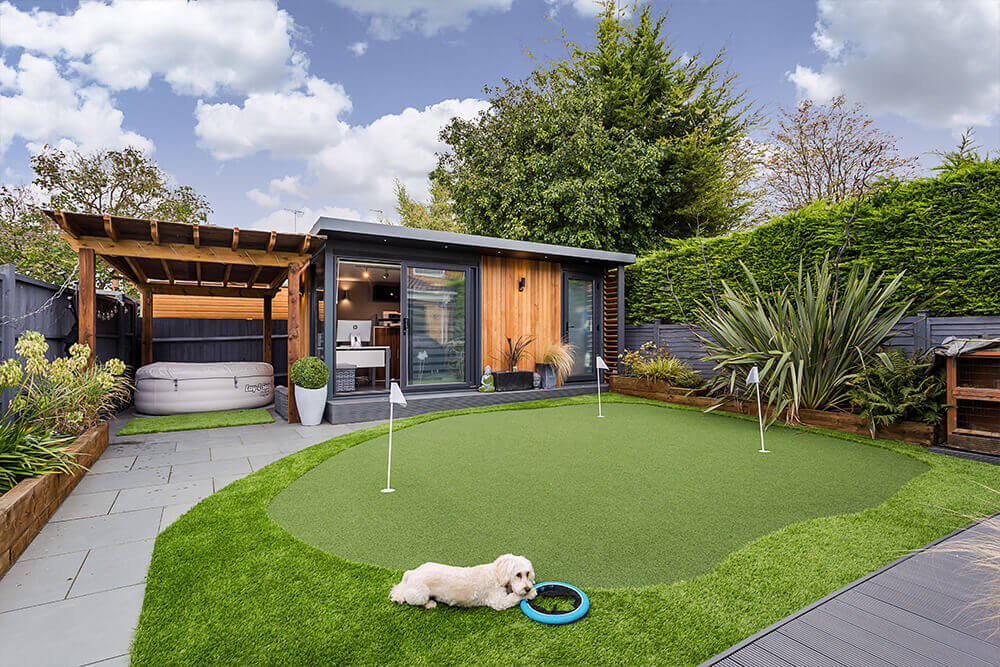 Golf astro putting green with small cream dog laying on it with wooden clad garden room.