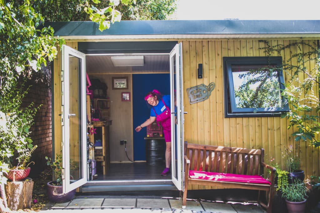 Exterior of a garden workshop home business with its doors open and with a bench and pink seat cushion. Our client is peeping happily from inside.