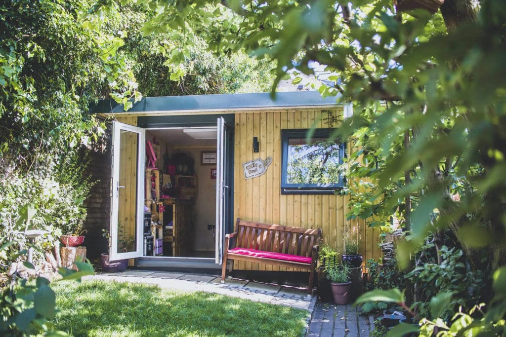 Exterior of a garden workshop home business with its doors open and with a bench and pink seat cushion.