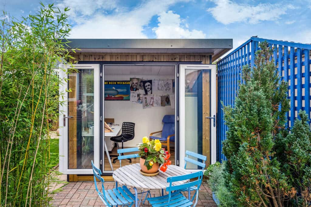 Wooden table and blue metal chairs in front of an open garden studio with its french doors open wide.
