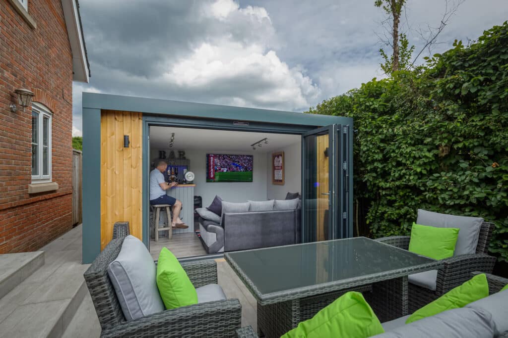 Exterior of Inspiration with bi-folds open looking into man cave with man on bar stool and garden furniture in front of the building