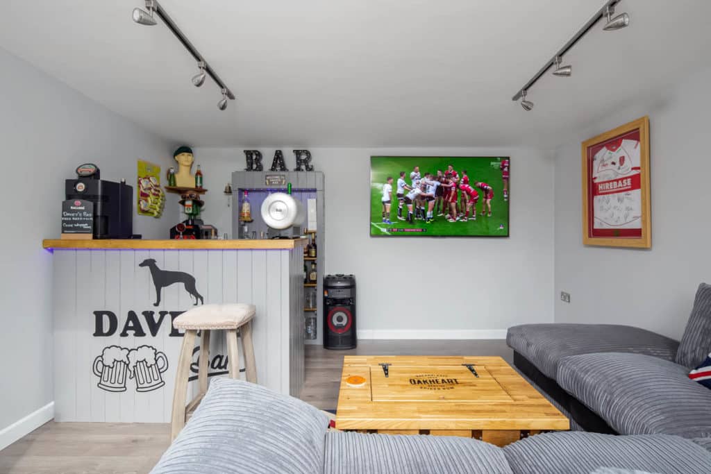 'Dave's bar' garden room interior with tv mounted on the wall in the background. B A R decorative letters sit on top of shelving unit with drinks displayed.
