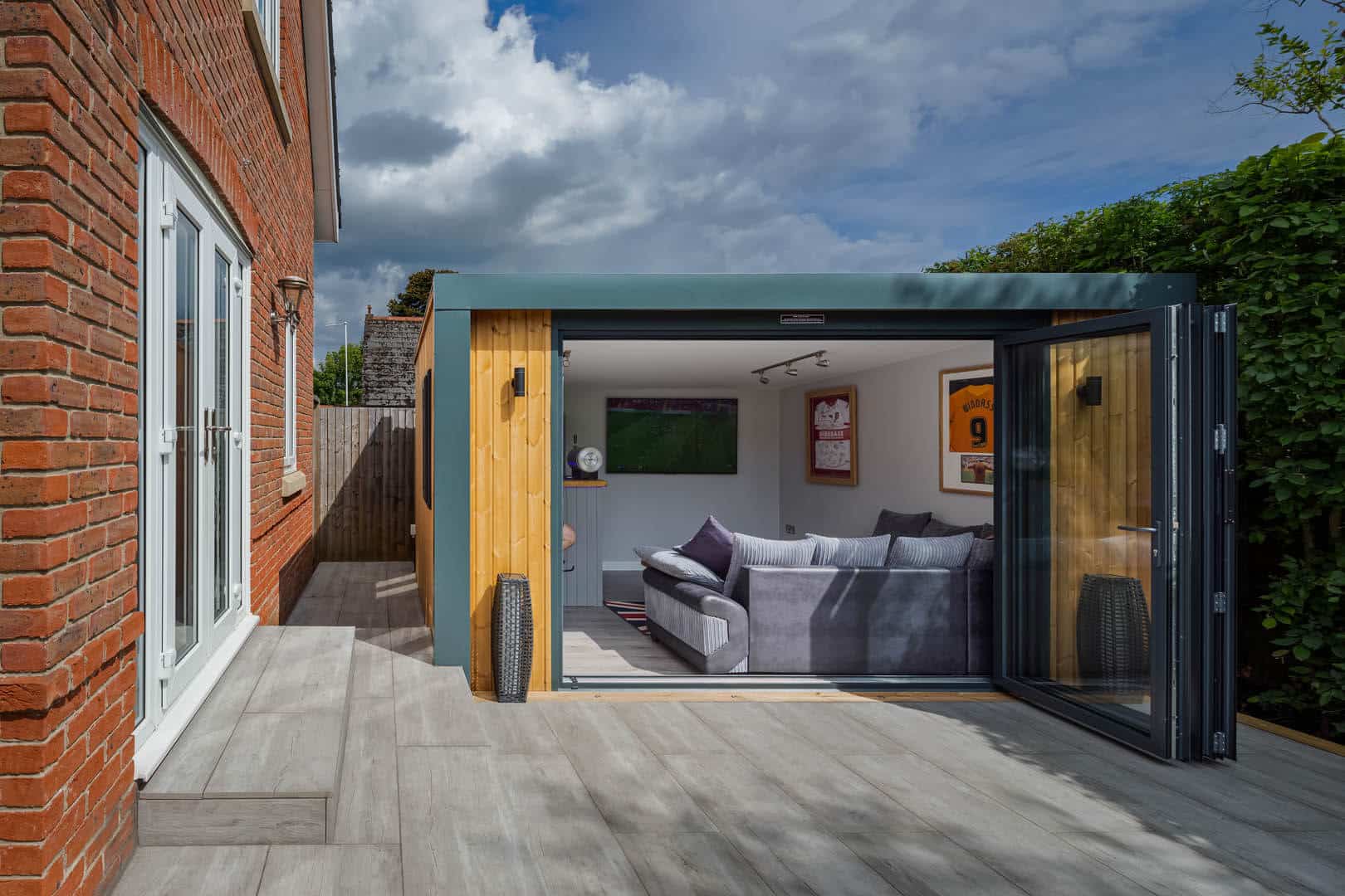 Exterior of a man cave on a patio. The bi-fold doors are open looking into the man cave with a grey L shaped sofa and TV mounted to the wall