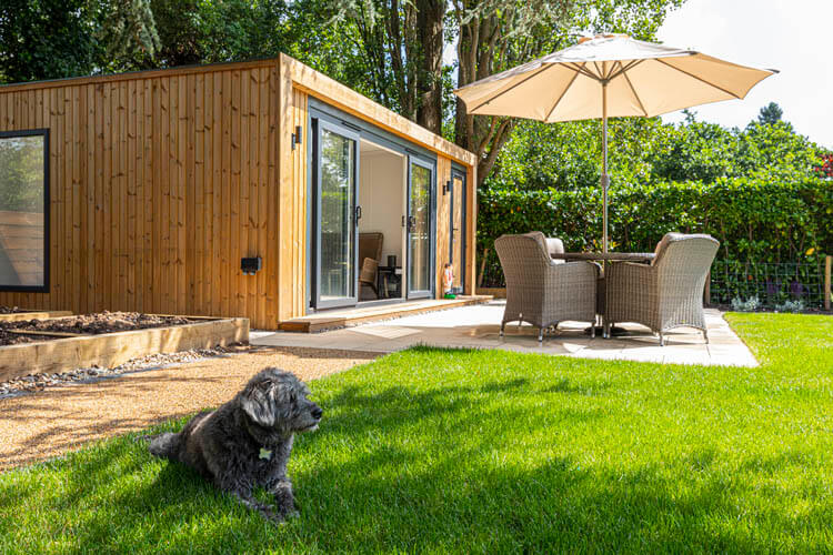 Garden with a path leading to a garden building with patio furniture outside and a parasol and a grey dog