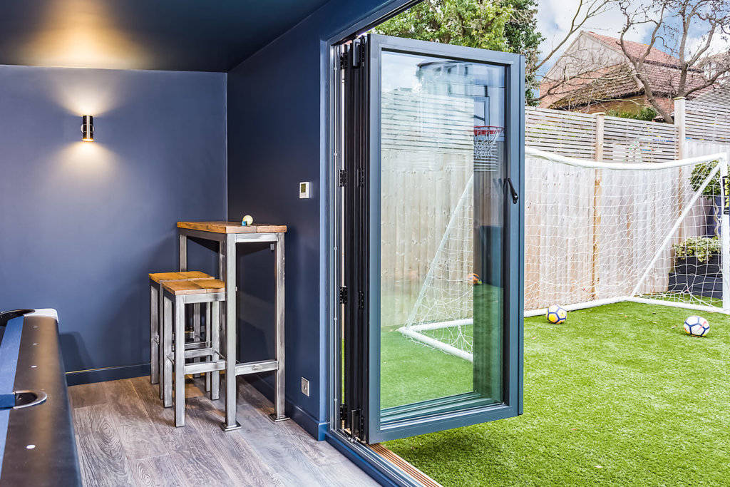 Dark blue interior of garden family game room with bifold doors open onto the garden lawn