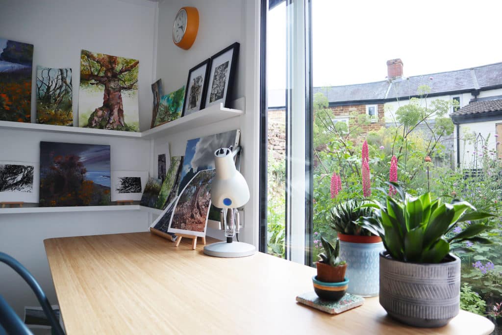 Inside garden room art studio of paintings on a shelf and plants on the desk