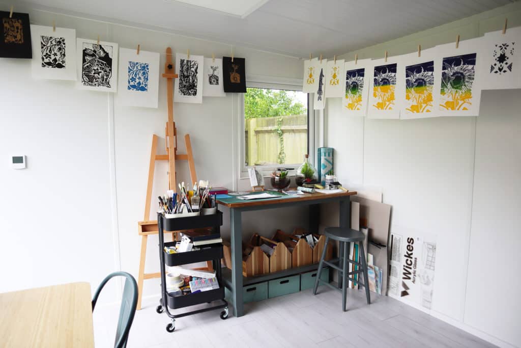 Inside garden room art studio with screen printing A4 samples handing on string and a work table filled with brushes and art utensils