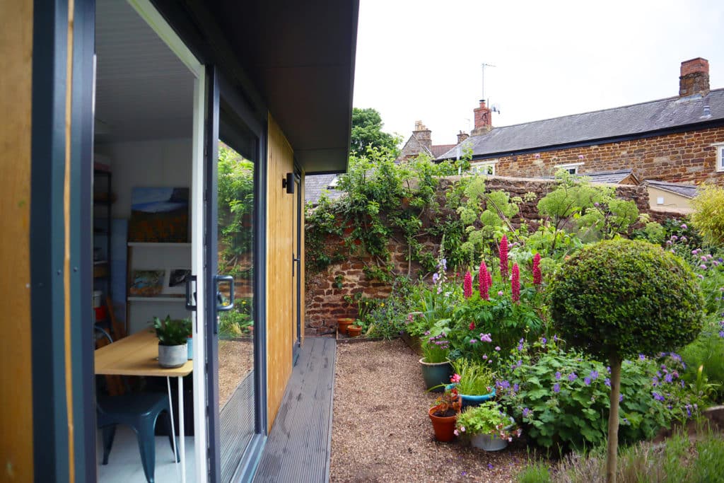 Side view of garden room peeking inside art studio and showing parts of the flower bed with multi coloured flowers