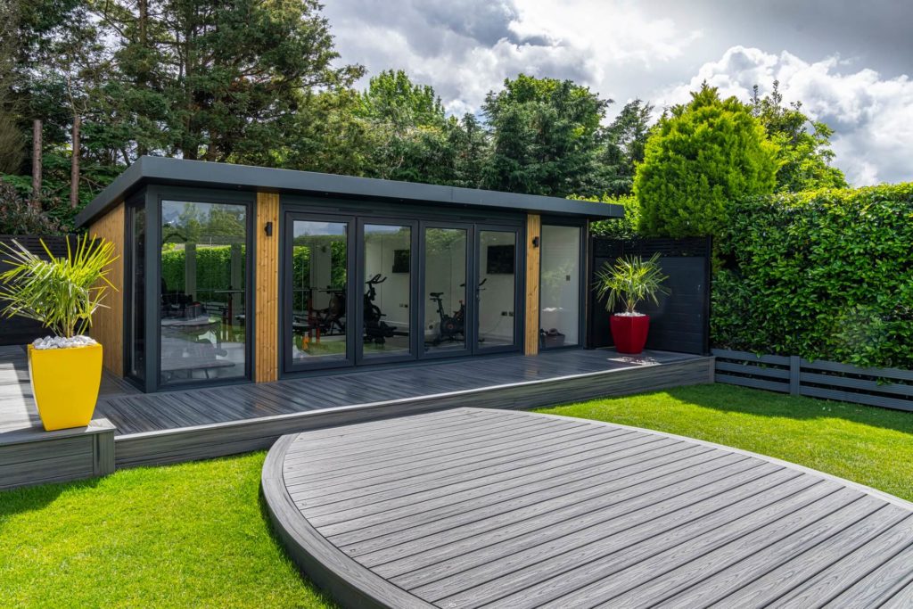 Garden room positioned on grey composite decking showing side angle and cladding. Large red and yellow plant pots also sit on the decking.