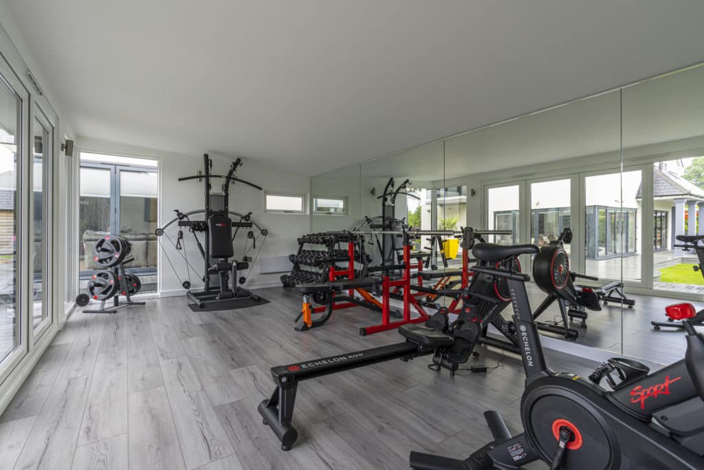 Full view of garden room gym equipment that is red and black. Back wall covered in mirrors reflecting outside garden