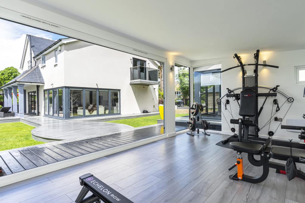 Gym equipment inside garden room gym with doors open looking back towards white rendered house