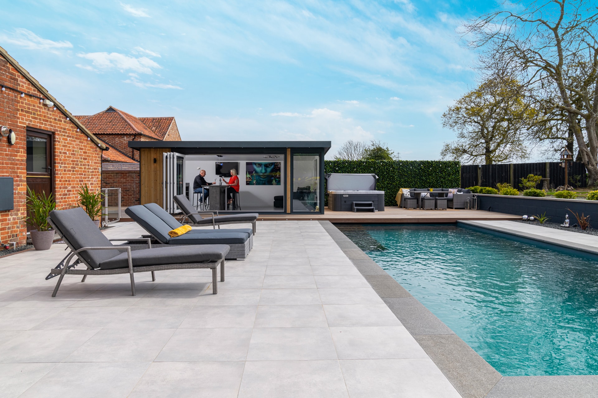Modern swimming pool surrounded by light grey paving with sun loungers on. Garden bar with customers in background.