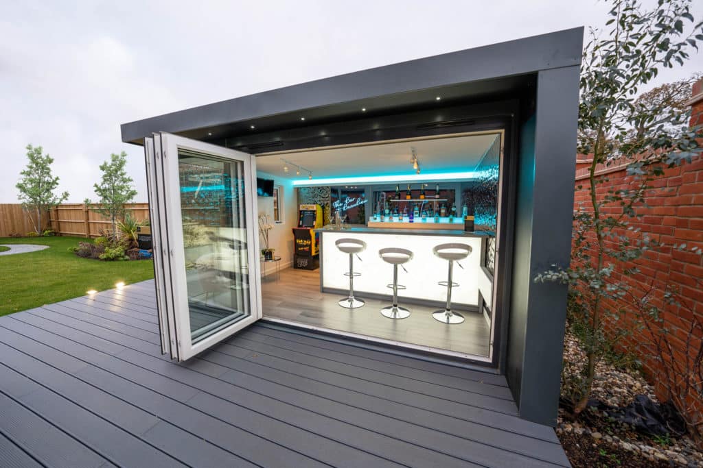 Garden room exterior with bifold doors open showing inside dressed as a garden bar with 3 bar stools, pac man machine and alcohol bottles displayed on shelf in background.