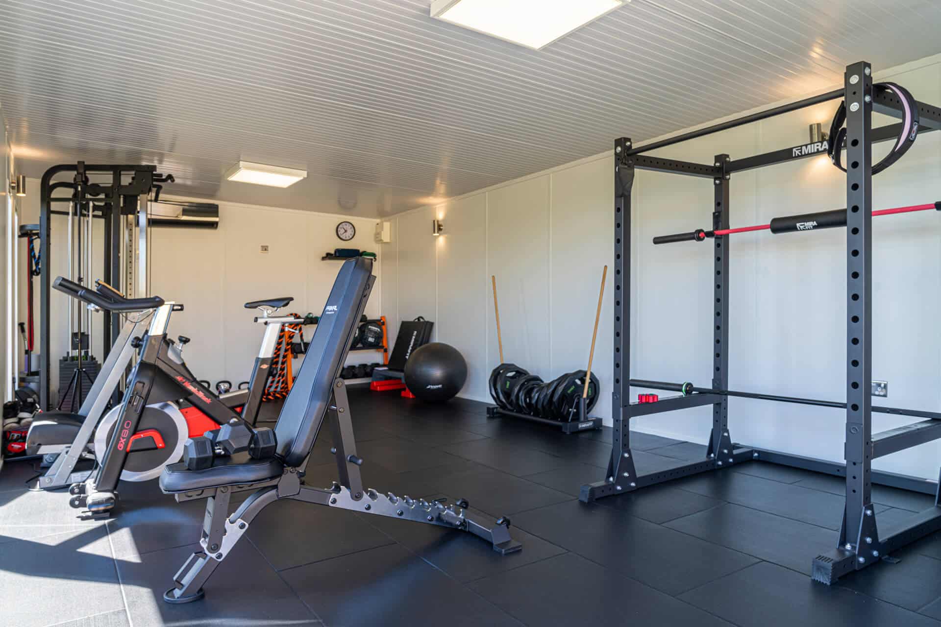 Gym equipment including weights, t-bar and bike positioned in home gym that has white interior walls.