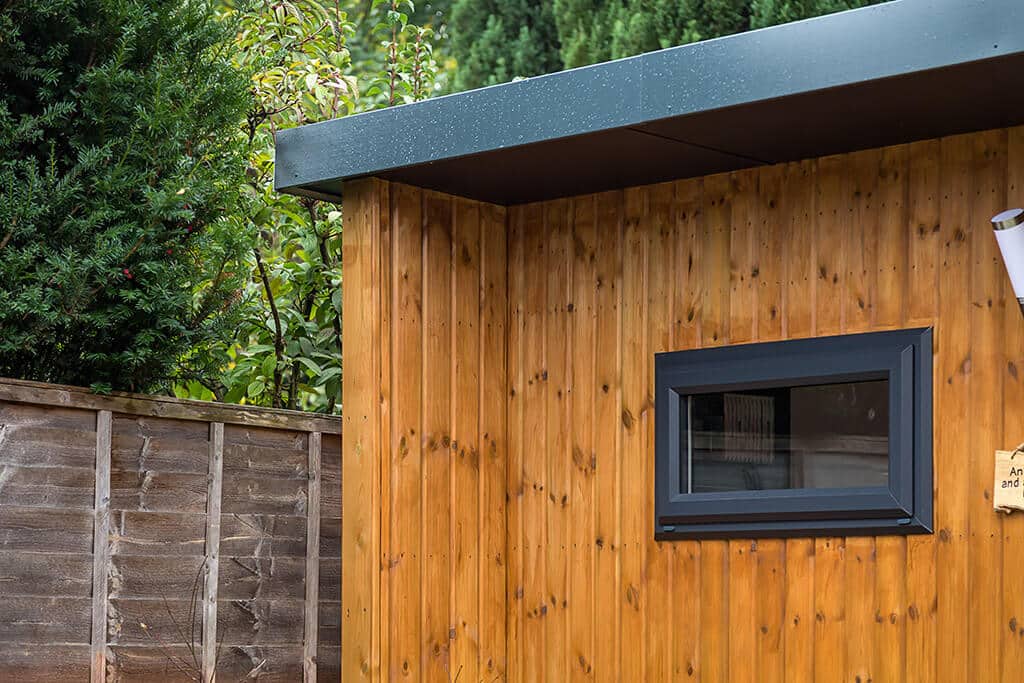 Close up of redwood clad garden room corner with graphite picture window and roof trim.