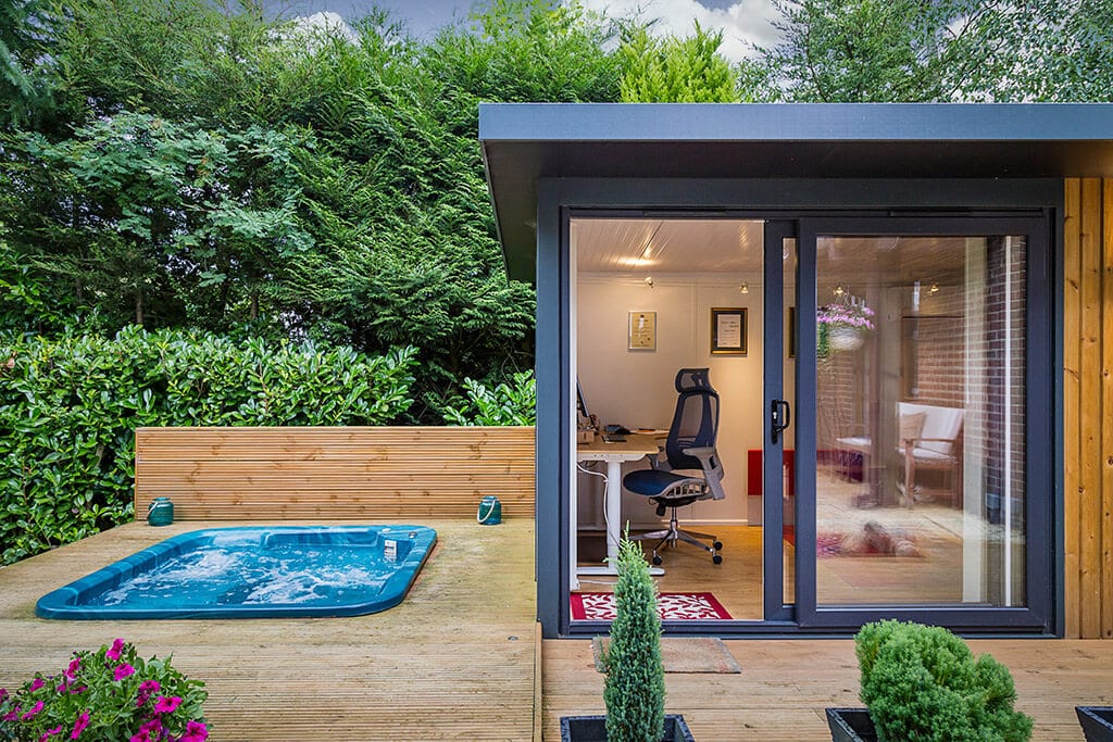 Sunken hot tub in decking with garden office with sliding graphite door open with view of office desk and chair.